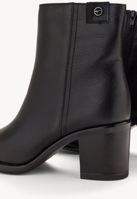 Černé kožené kotníkové boty s texturovaným povrchem, blokovým podpatkem, bočním zipem a dekorativním logem na zadní straně.