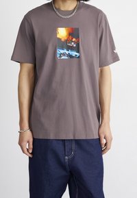 Grå bomullströja med en central grafisk tryck i livliga färger. Kortärmad och med en avslappnad passform, bärs tillsammans med blå denimbyxor.