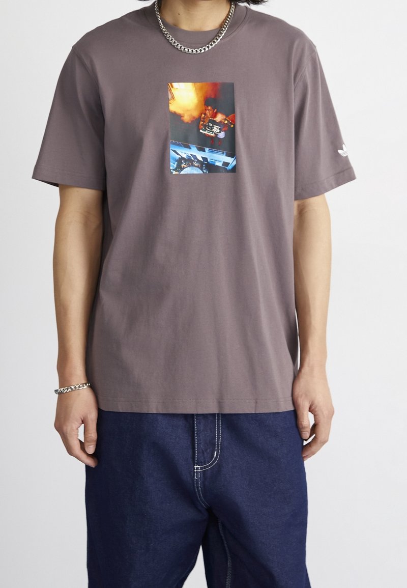 Grå bomullströja med en central grafisk tryck i livliga färger. Kortärmad och med en avslappnad passform, bärs tillsammans med blå denimbyxor.