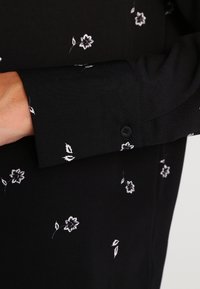 Blouse noire présentant un imprimé floral blanc, avec un détail de manchette boutonnée. Le tissu a une texture lisse et un léger brillant.