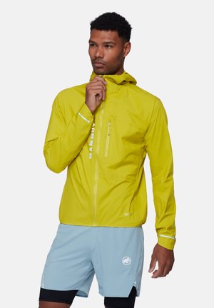 Mann in gelber Kapuzenjacke und hellblauen Shorts, der den Reißverschluss der Jacke anpasst und vor einem neutralen Hintergrund steht.