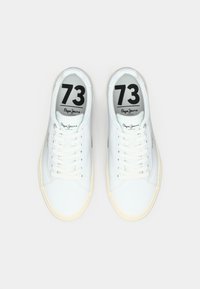 White leather sneakers with round toes, flat laces, and beige rubber soles. Black printed logo and number '73' on insoles.