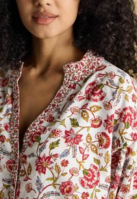 Kwiatowa bawełniana bluzka o białym tle z czerwonymi, zielonymi i niebieskimi wzorami. Zawiera zapięcie na guziki i marszczony dekolt.