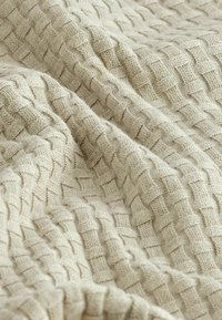 Tissu en tricot beige avec un motif tissé texturé présentant des carrés surélevés créant de la profondeur. Texture douce et chaude parfaite pour des couvertures ou des vêtements.