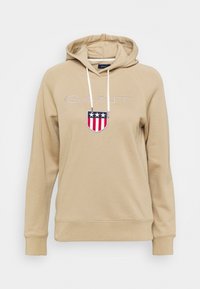 Felpa con cappuccio beige con logo "GANT", uno scudo con strisce rosse e blu, e cordini bianchi. Tessuto morbido, polsini e orlo a coste.