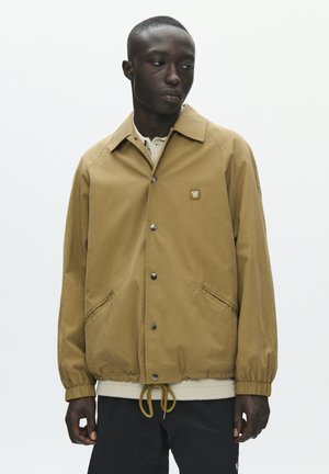 Mand iført lys brun, knappet jakke med elastiske manchetter og snørre i kanten, stående foran en ensfarvet hvid baggrund.