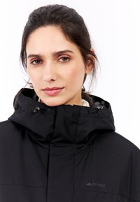 Sort hættejakke med høj krave, der har snore og blødt foer. Diskret logo på brystet. Vandtæt materiale.