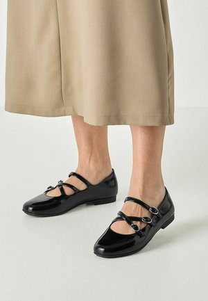 Fødder iført sorte laklæder Mary Jane-sko med to spænder, beige bukser med vide ben, der er kortet af ved midt på læggen.