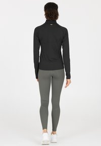 Zwarte long sleeve top met hoge kraag, gecombineerd met lichtgrijze geribde leggings. Model draagt witte sneakers en staat met de rug naar de camera.