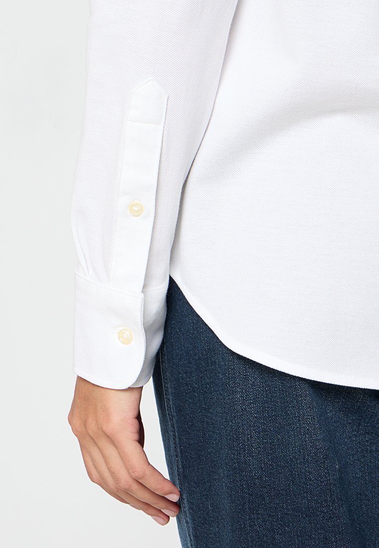 Camicia bianca a maniche lunghe con una finitura testurizzata, dotata di due bottoni giallo chiaro sui polsini e un orlo arrotondato, abbinata a jeans blu scuro.