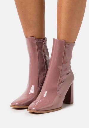 Glanzende mauve enkellaarsjes met blokhakken en zijritsen, gedragen op blote benen, afgebeeld tegen een witte achtergrond.