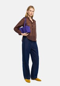 Brązowa przezroczysta koszula z guzikami, niebieskie dżinsy o szerokich nogawkach oraz żółte zamszowe buty, trzymająca małą fioletową torebkę z materiału.