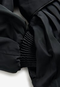 Černá bunda s plisovaným designem; má elastické manžety a hladkou texturovanou tkaninu. Jemný lesk dodává hloubku.