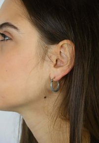 Srebrna kolczyk-koło na uchu modelki, z teksturowanym wzorem o okrągłym kształcie i cienkimi, plecionymi detalami.