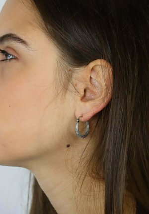 Silberner Creolen-Ohrring, der am Ohr eines Modells präsentiert wird. Er zeigt ein strukturiertes Design mit runder Form und feinen, geflochtenen Details.