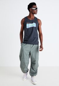 Svart ermeløs topp med "Greetings from Levi's" grafikk, kombinert med løse, lys grønne cargo-bukser og lavendel joggesko.