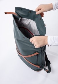 Zaino impermeabile verde con dettagli in pelle marrone; presenta una parte superiore aperta, tasca interna con zip e foderato a scacchi; spallacci regolabili.