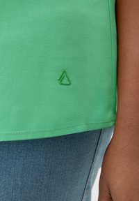 Zelena majica od glatkog materijala s izvezenim zelenim trokutastim logotipom na rubu, u kombinaciji s plavim trapericama.