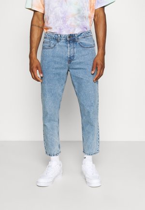 Homme portant un jean bleu clair court, des baskets blanches, des chaussettes blanches et un t-shirt tie-dye pastel, debout devant un fond uni.
