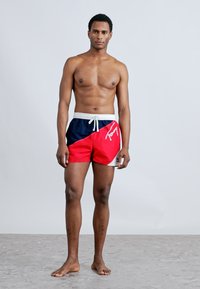 Ανδρικό μαγιό σορτς σε ναυτικό μπλε και κόκκινο, με λευκό κορδόνι στη μέση, λογότυπο και αθλητική σχεδίαση με γεωμετρικό χρωματικό σχέδιο.