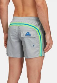 Una persona sta in piedi con le spalle alla fotocamera, indossando pantaloni da bagno grigi con strisce laterali blu e verde neon e una tasca applicata.