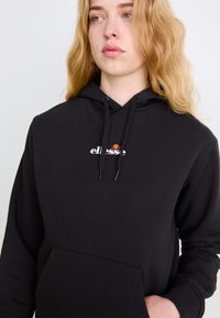 Sudadera negra con bolsillo delantero, cordones ajustables y un logo blanco. Textura de tela suave y ajuste relajado.