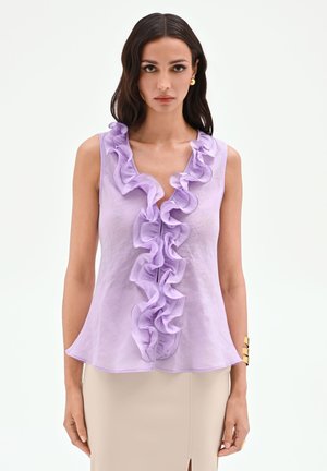 Femme portant un chemisier lilas sans manches à volant sur le devant et une jupe beige, debout devant un fond clair uni.