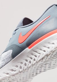 Gros plan de la semelle grise et blanche d'une chaussure de course avec le swoosh orange de Nike et un motif détaillé de la bande de roulement sur fond blanc.