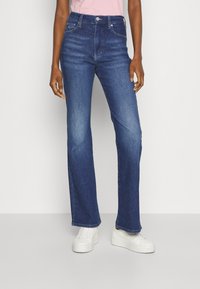 Jeans de mezclilla azul oscuro con un diseño de pierna recta, cintura alta y un sutil desgaste en las rodillas. Combinados con zapatillas blancas.