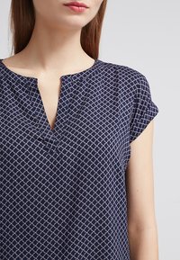 Námořnická modrá košile s krátkými ohrnutými rukávy, s diamantovým vzorem v světle modré. V-krkový design s malým otevřením na výstřihu.