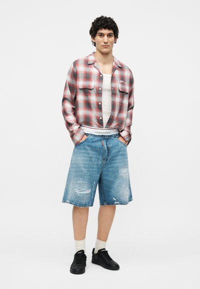 Νέος άνδρας στέκεται με τα χέρια στις τσέπες φορώντας κόκκινο καρό πουκάμισο, λευκό αμάνικο, σκισμένο denim σορτς, λευκές κάλτσες και μαύρα αθλητικά παπούτσια.