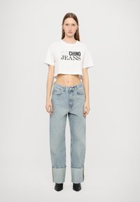 Balta trumpa marškinėliai su juodais užrašais "CHINO JEANS"; šviesiai mėlynos, plačios kojos džinsai su sulankstytomis rankovėmis; juodi batai.