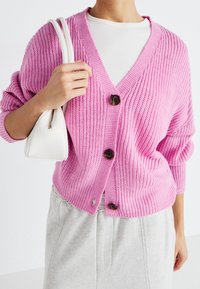 Rosa stickad cardigan med V-halsringning, med tre stora bruna knappar och avslappnade ärmar. Matchad med en ljusgrå outfit och en vit väska.