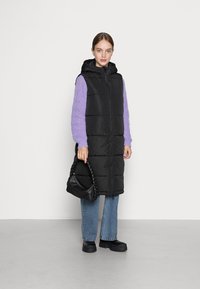 Padded long vest noir avec une capuche, porté sur un pull en grosse maille violet clair. Le mannequin tient un sac avec une anse tressée noire. Style décontracté en denim.