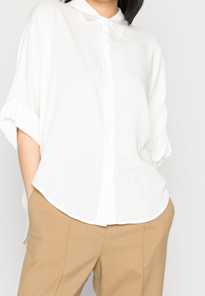 Femme portant une chemise blanche ample boutonnée avec les manches retroussées et un pantalon beige, mains dans les poches, debout devant un fond uni.