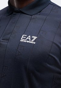 Gros plan d'un polo bleu marine foncé avec un col, arborant le logo blanc EA7 Emporio Armani sur la poitrine.