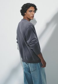 Pelēks, pārmērīgs svītrojums ar garām piedurknēm, ribotas aproces un apakšmalu; valkāts ar gaiši ziliem, brīvi pieguļošiem džinsiem uz vienkārša baltā fona.