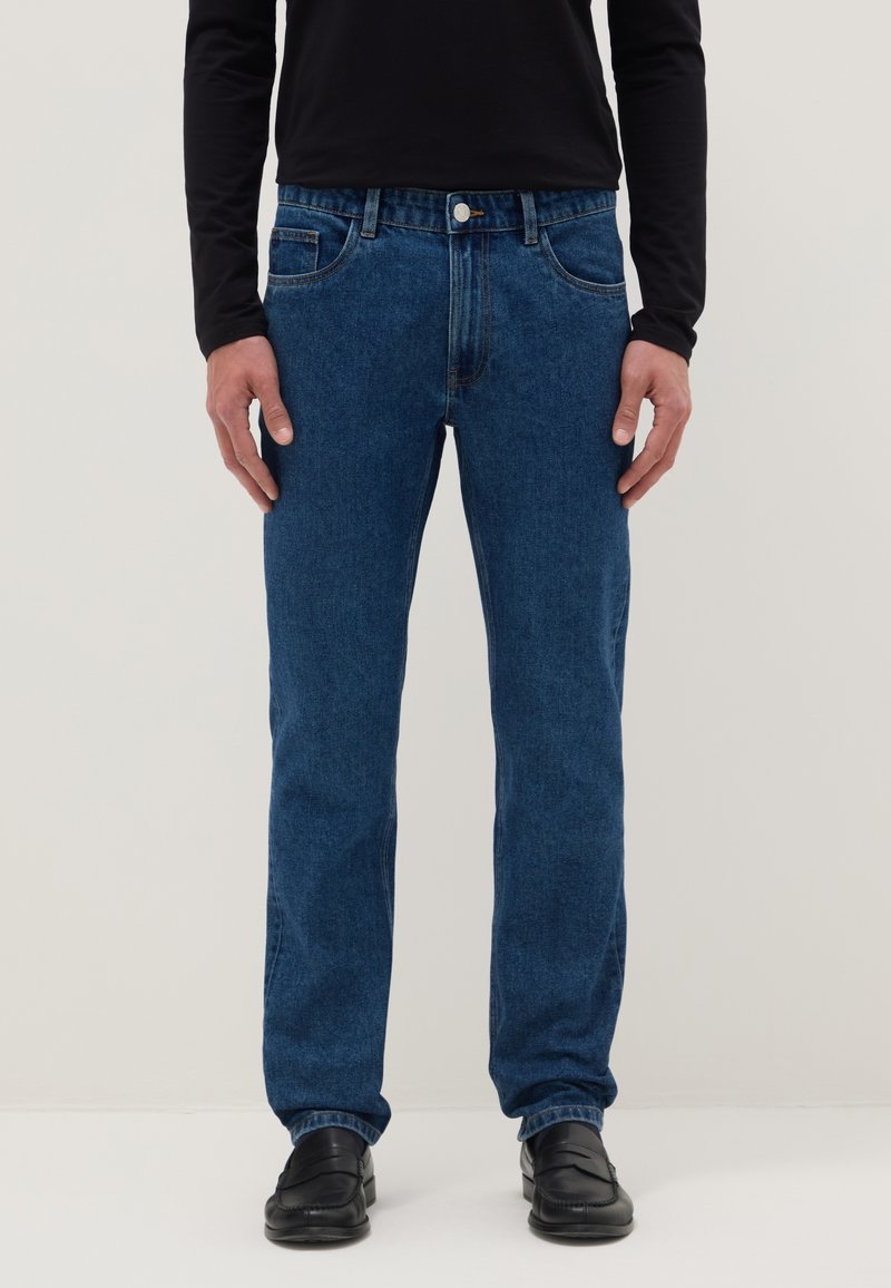 Hombre vistiendo jeans azules de corte recto, camisa negra de manga larga y mocasines negros, posando frente a un fondo claro y liso.