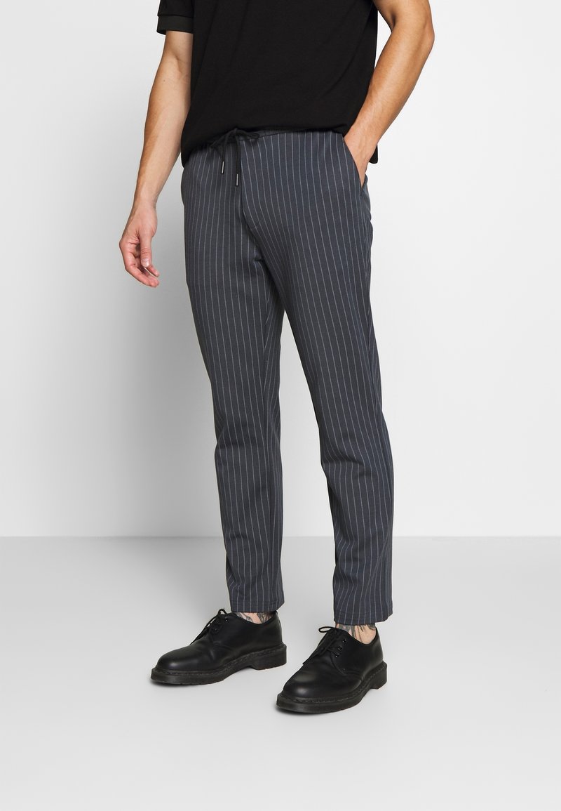 Homme portant un pantalon gris foncé à fines rayures avec taille à cordon et des chaussures en cuir noires à lacets, debout avec une main dans la poche.