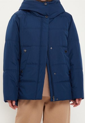 Personne portant une veste matelassée bleu marine avec des pressions et une fermeture éclair, associée à un pantalon beige, sur un fond uni.