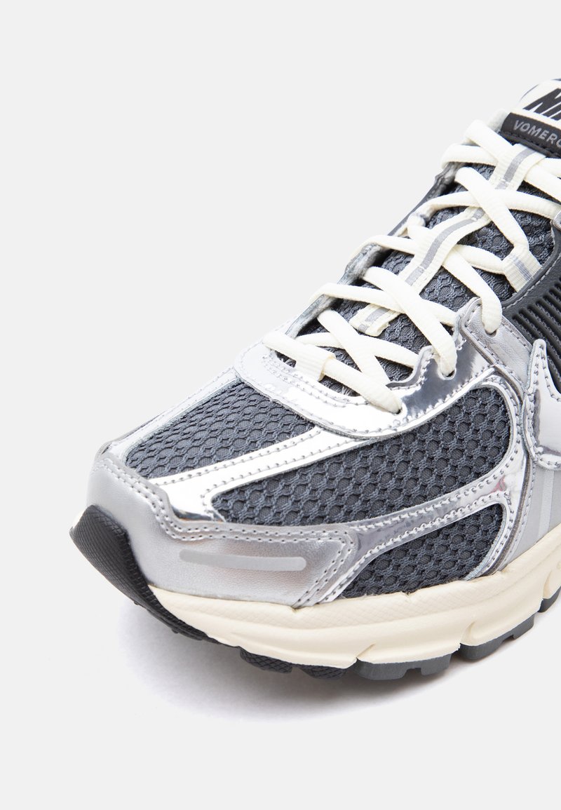 Zapatilla de running Nike en gris oscuro y plateado con cordones blancos y parte superior de malla texturizada, vista desde un ángulo frontal lateral sobre fondo blanco.