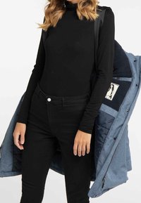 Maglietta nera a maniche lunghe abbinata a jeans neri, con una giacca di jeans blu in mano. La giacca presenta un interno trapuntato e accenti di tessuto a contrasto.