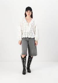 White eyelet blouse with ruffled sleeves, tied front. Grey denim shorts. Black knee-high boots with intricate white embroidery. Black cap.