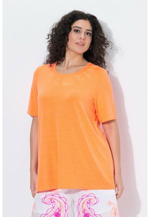 Kvinde med krøllet hår iført en orange T-shirt med korte ærmer med muslingeskaldesign og hvide bukser med lyserøde paisleymønstre, stående mod hvid baggrund.