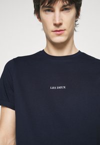 Tamsiai mėlynas apvalios kaklo marškinėliai, pagaminti iš medvilnės, su trumpomis rankovėmis ir baltu „LES DEUX“ logotipu, esančiu centrinėje krūtinės dalyje.
