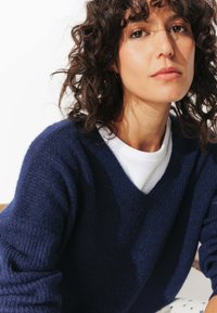 Un pull bleu marine en V, fabriqué en matériau doux avec une finition texturée, superposé sur une chemise blanche à col rond. Présente des manches larges.