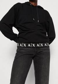 Černá mikina s kapucí se strukturovaným povrchem, s bílým značkovým elastickým pásem na spodním lemu, spárovaná s tmavými denimovými džíny.