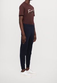 Marooninpunainen puuvillainen t-paita, jossa on vaaleanvärinen "EAT"-grafiikka, yhdistetty tummansinisiin collegehousuihin ja valkoisiin lenkkareihin, joissa on vihreitä yksityiskohtia, sivukuva.