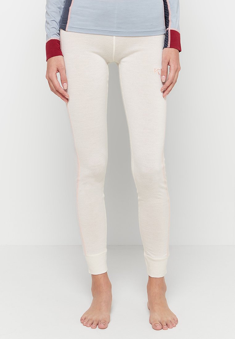 Femme portant des leggings ajustés de couleur crème et un haut à manches longues avec des manches rouges et bleues, debout pieds nus sur un sol blanc.