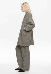 Mujer rubia con chaqueta de traje de gran tamaño y pantalones de pierna ancha, ambos con patrón de pata de gallo en blanco y negro, de pie de perfil con las manos en los bolsillos.